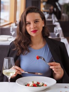 Řecká žena jí řecký salát s černým sakem a šumivým vínem. Červené rty a modré tričko.